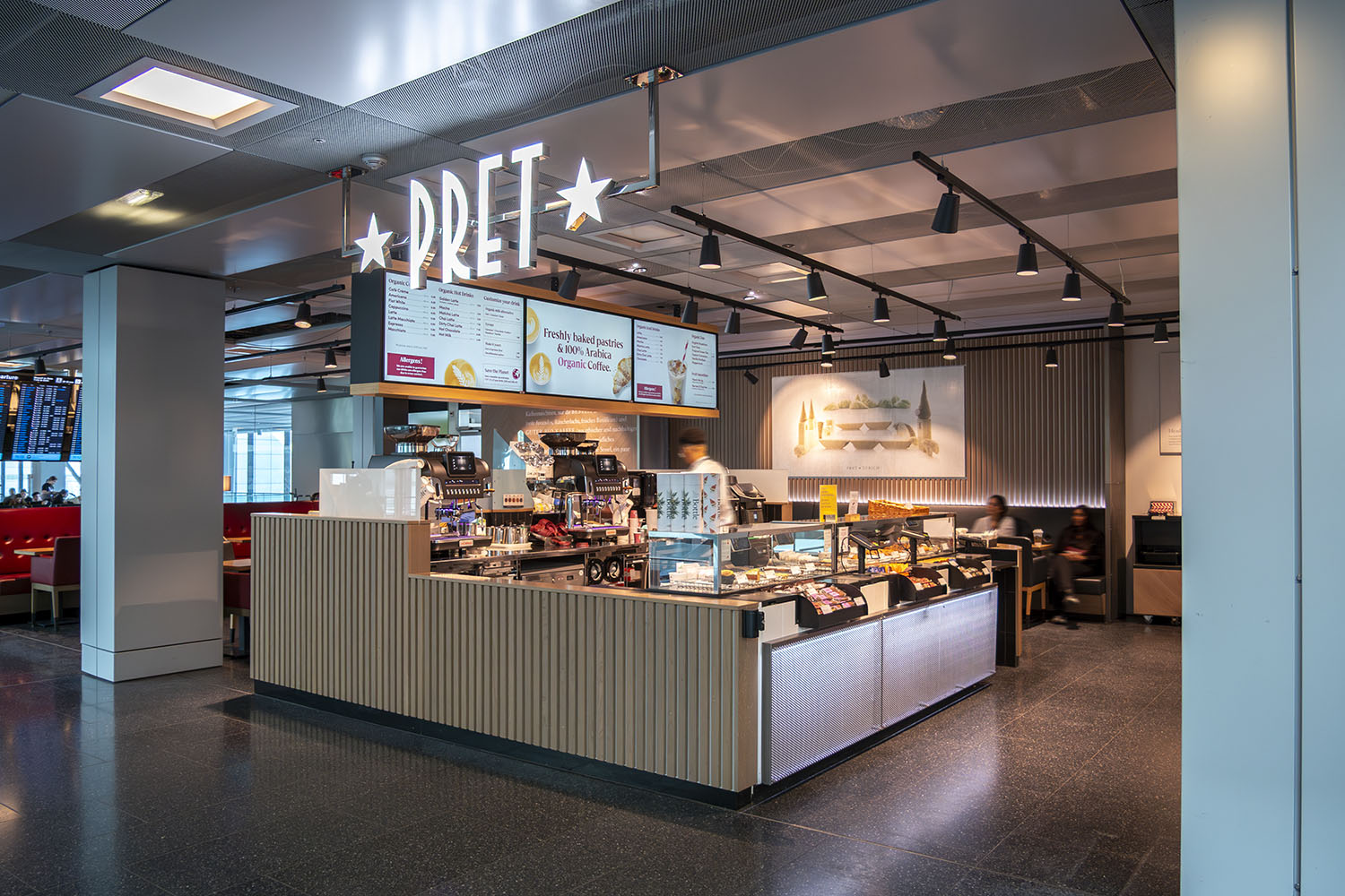 Moderne Pret Cafétheke mit beleuchtetem Logo, Kaffeemaschinen und einer Vitrine mit Backwaren in einem Flughafen.