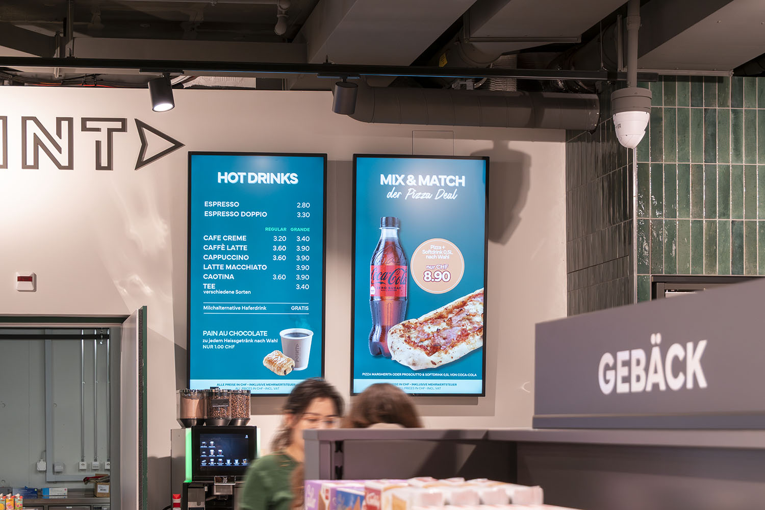 Digitale Menütafeln zeigen heiße Getränkepreise und ein Pizza-Softdrink-Angebot für 8,90 CHF in einem Café.