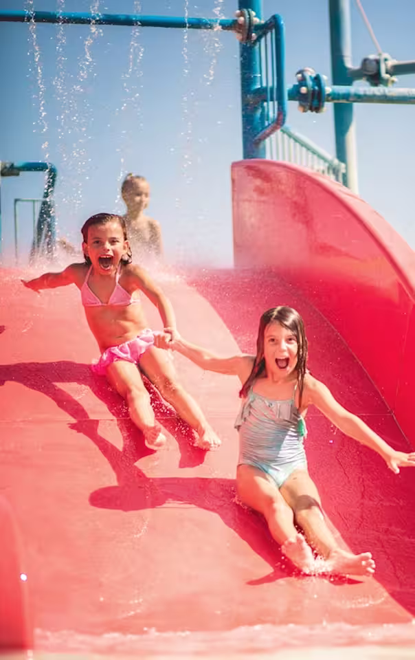 Two young girls in swimsuits holding hands and sliding down a red water slide with water spraying around them.