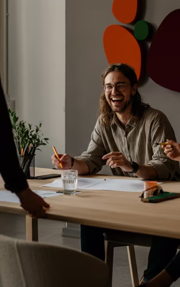 Man with glasses laughing and holding a pen while sitting at a table with papers and a glass of water.