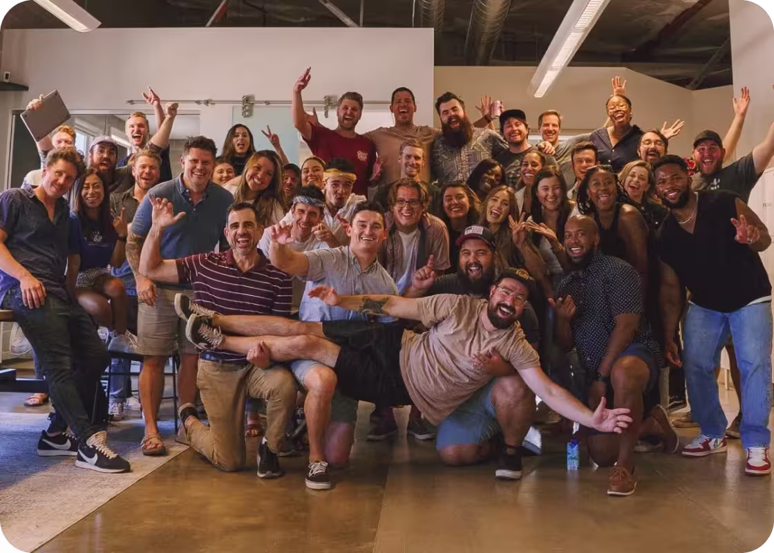 Large group of diverse people smiling and posing energetically indoors, with one man being playfully held up horizontally in front.