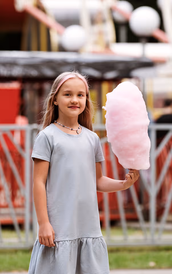 Young girl in a light gray dress holding large pink cotton candy at an outdoor fair.