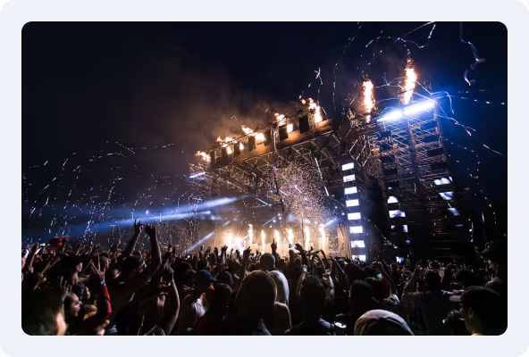 Crowd of people at an outdoor concert at night with a large stage lit by bright lights and pyrotechnics.