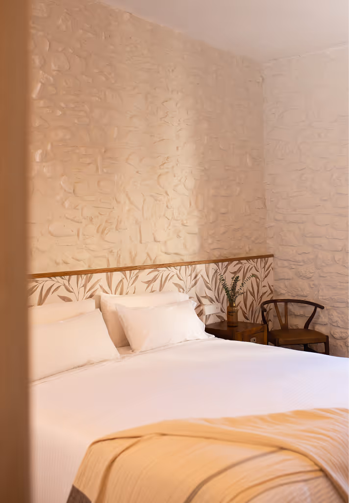 Cozy bedroom with white bedding, beige throw blanket, textured stone walls, a wooden chair, and a small bedside table with a vase of greenery.