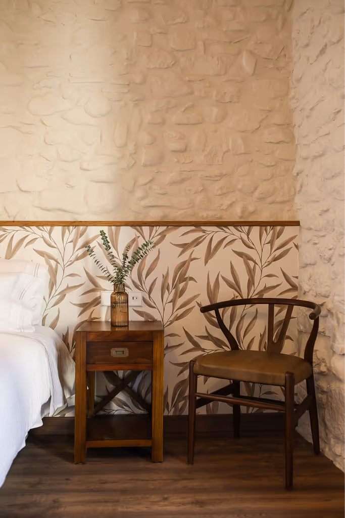 Wooden nightstand with a brown vase holding green leaves, next to a dark wooden chair and a bed with white linens against a textured beige wall with leaf-patterned wallpaper on the lower half.