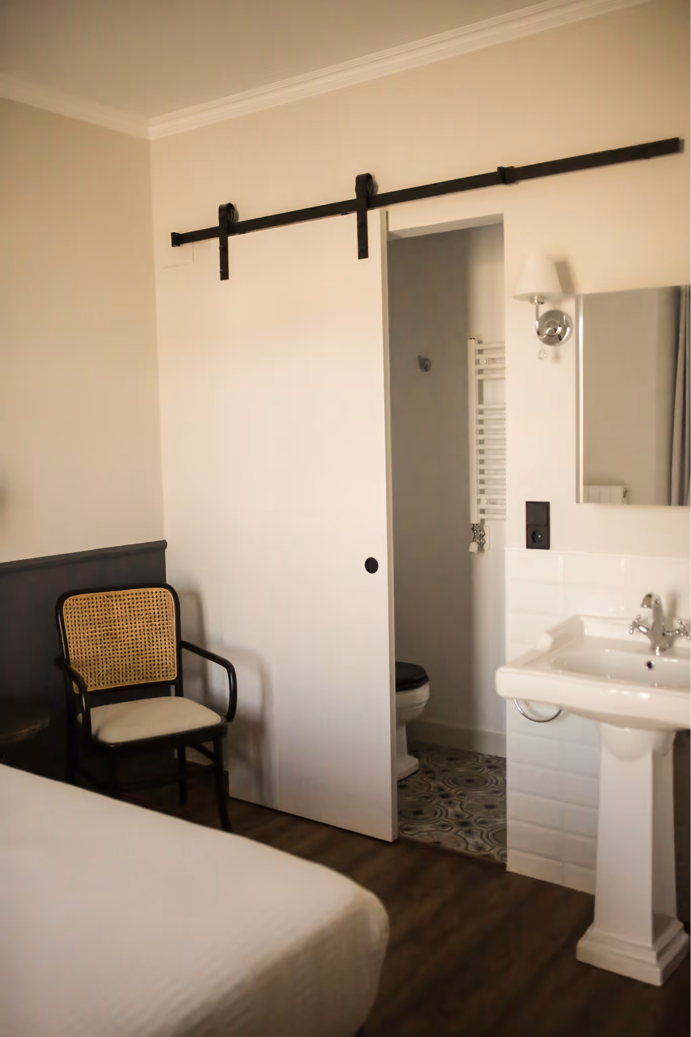 Minimalist bedroom corner with a wooden chair, white sliding barn door partially open to a bathroom, and a white pedestal sink with mirror and wall lamp.