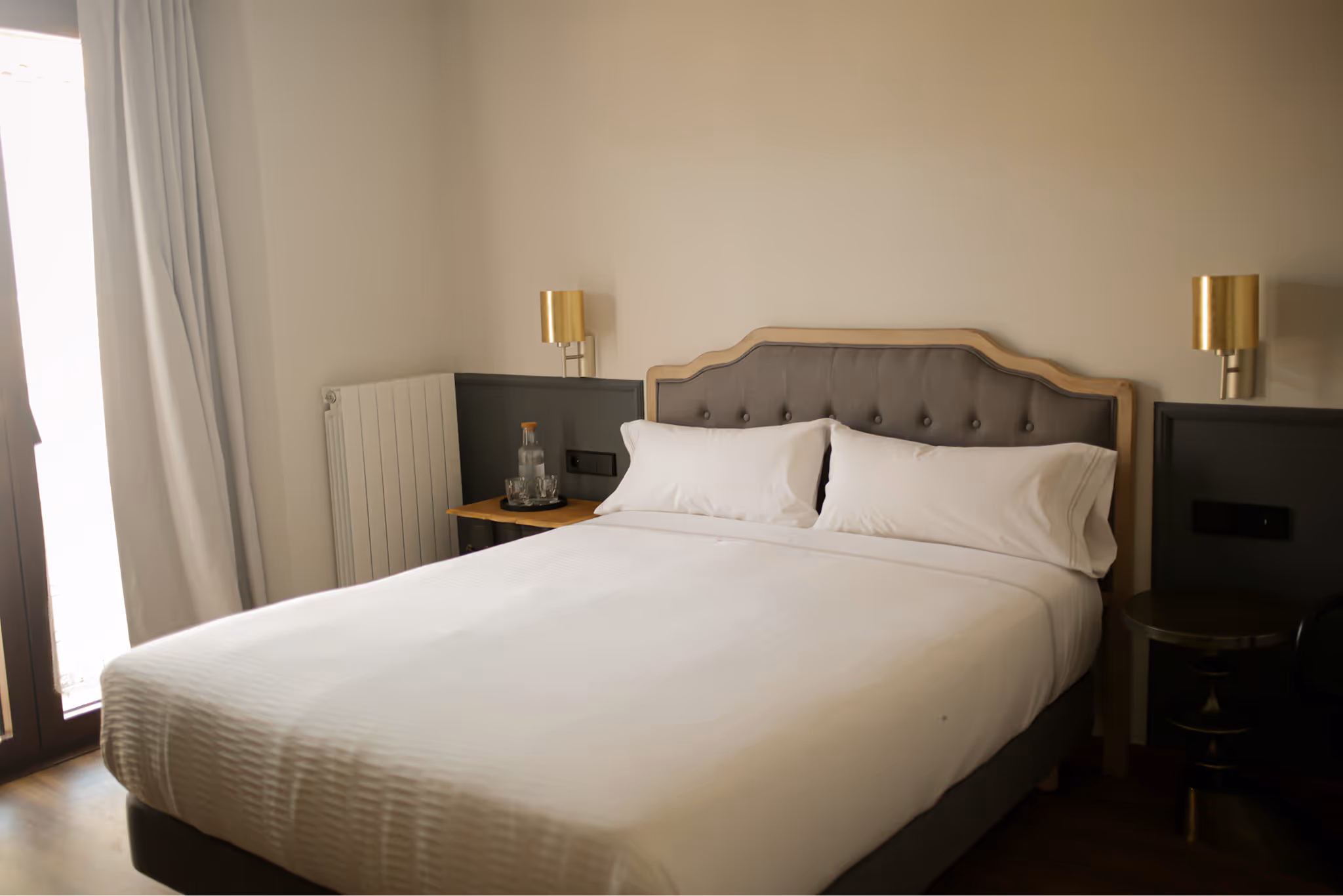 Neatly made bed with white linens and two pillows in a minimalist bedroom with beige walls, two gold wall lamps, and a small table holding a glass bottle and glasses.
