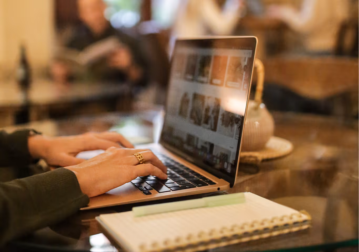 Imagen en primer plano de unas manos de mujer trabajando sobre un ordenador portátil. Al fondo se aprecian de forma borrosa personas en otras mesas de la estancia.