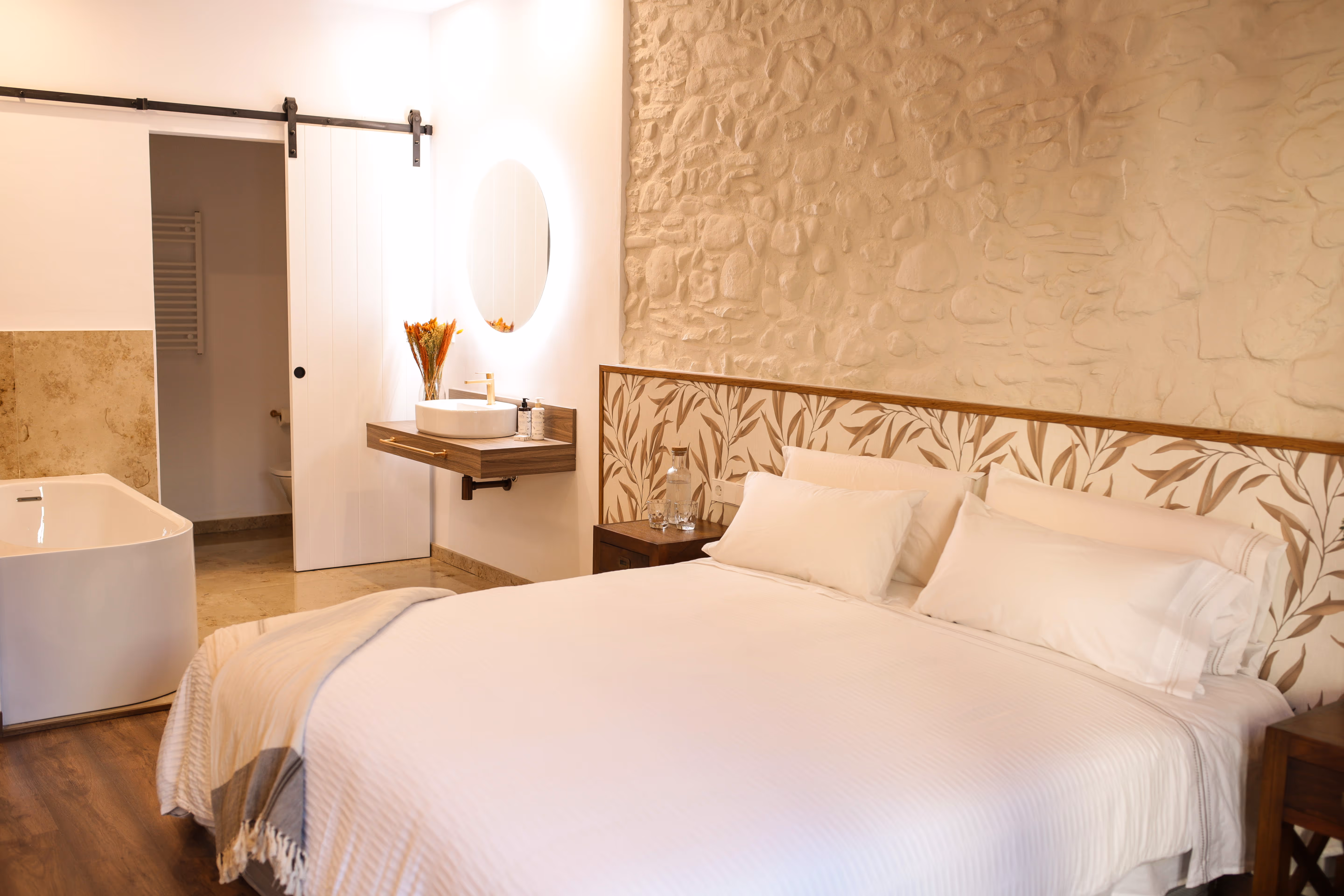Bedroom with a large white bed, patterned headboard, stone textured wall, floating sink with round mirror, bathtub, and sliding door to bathroom.
