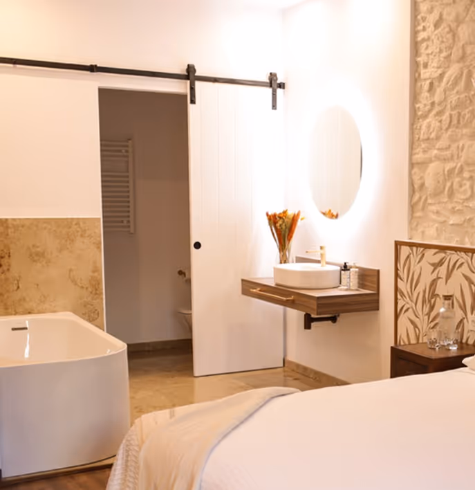 Modern bedroom with white bed, freestanding bathtub, floating sink with round illuminated mirror, and sliding barn door.