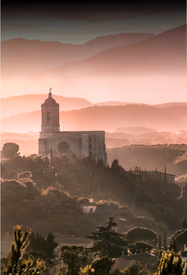 Historic stone church on a hill surrounded by trees with layered mountains in the background under a soft pinkish sky.