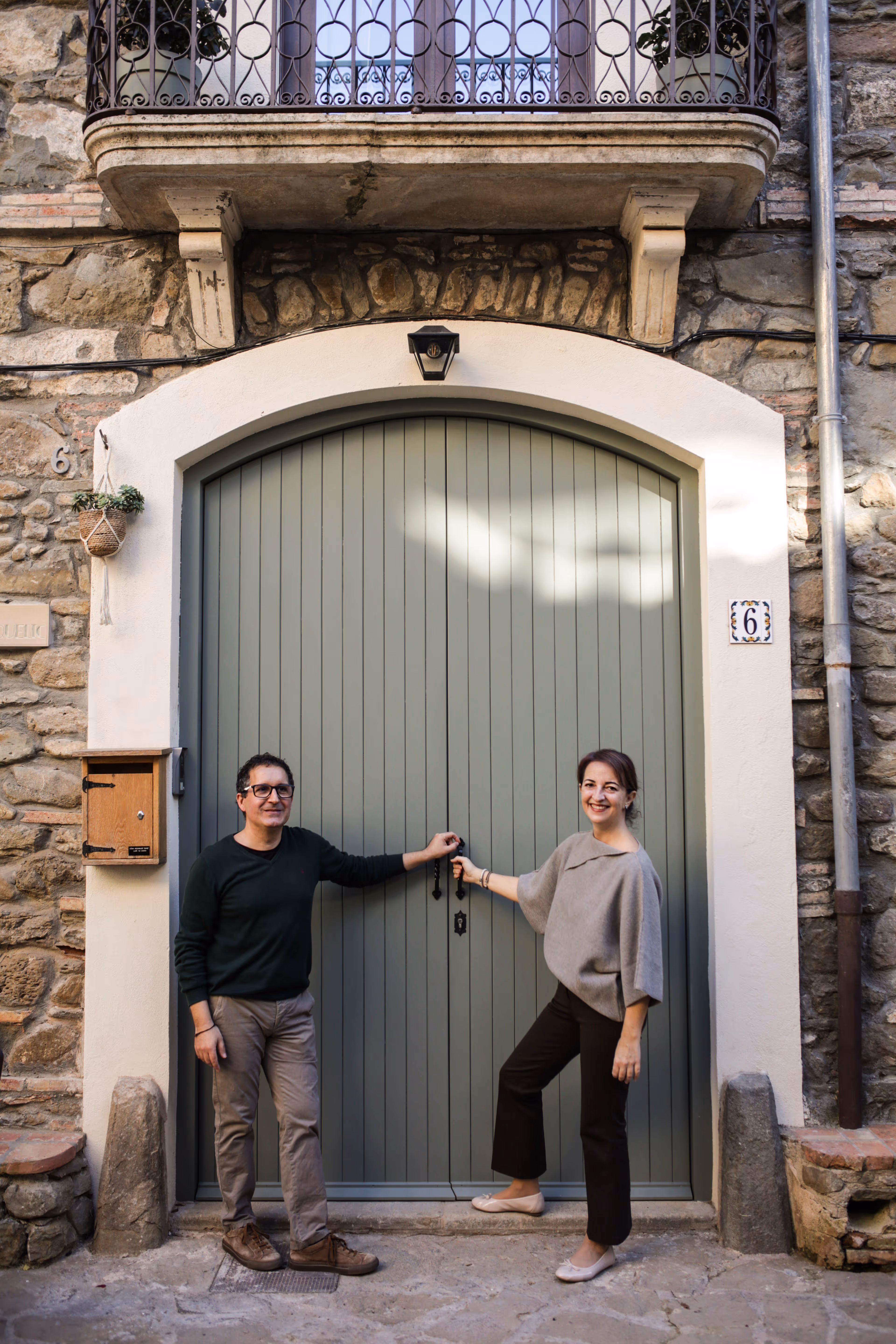 A man and woman standing in front of a large green arched door on a stone building, holding the door handles while smiling.