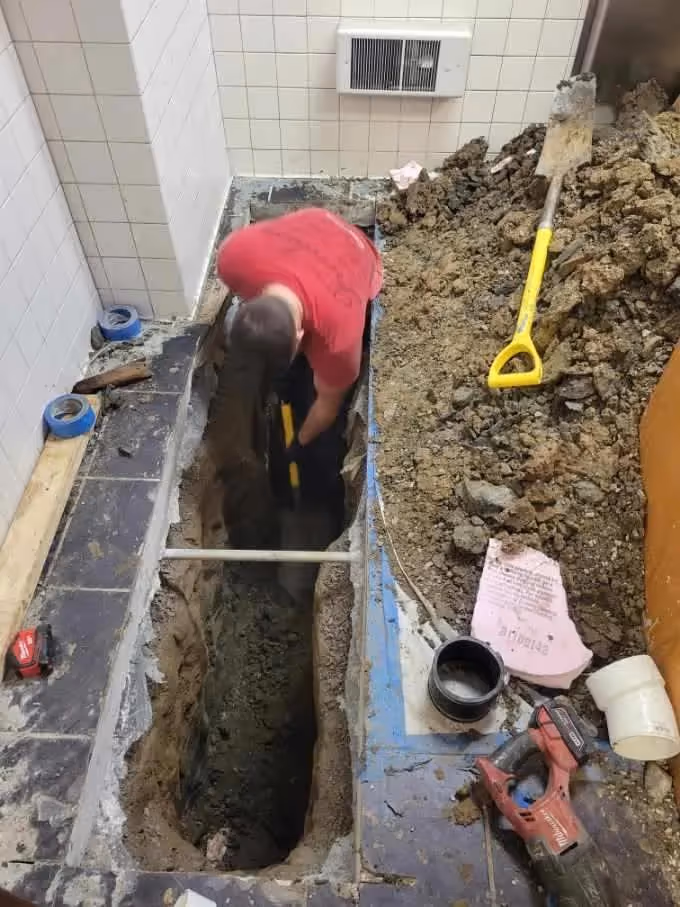 Technician digging a narrow trench around exposed pipes during a bathroom renovation