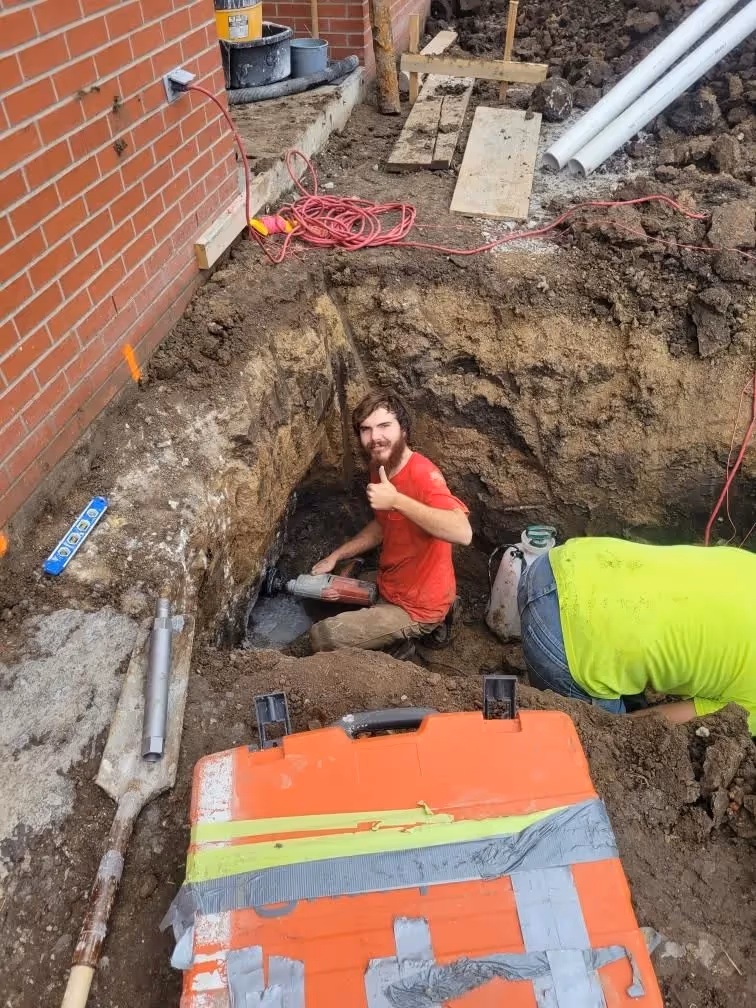 Worker in a red shirt inside a deep excavation near a brick wall installing pipes.