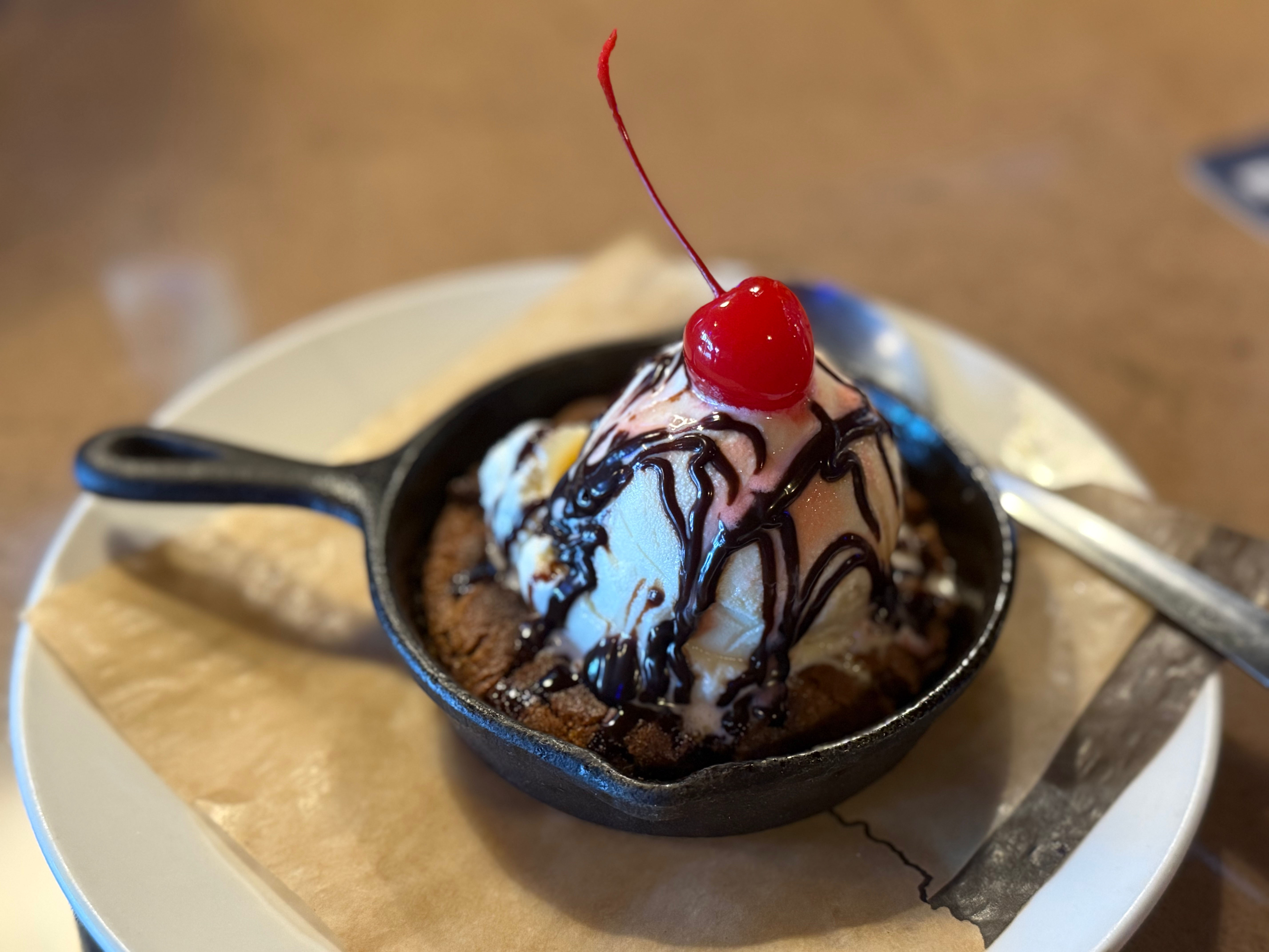 Skillet cookie topped with a scoop of vanilla ice cream, chocolate syrup, and a red cherry on top, served on a plate with a spoon.