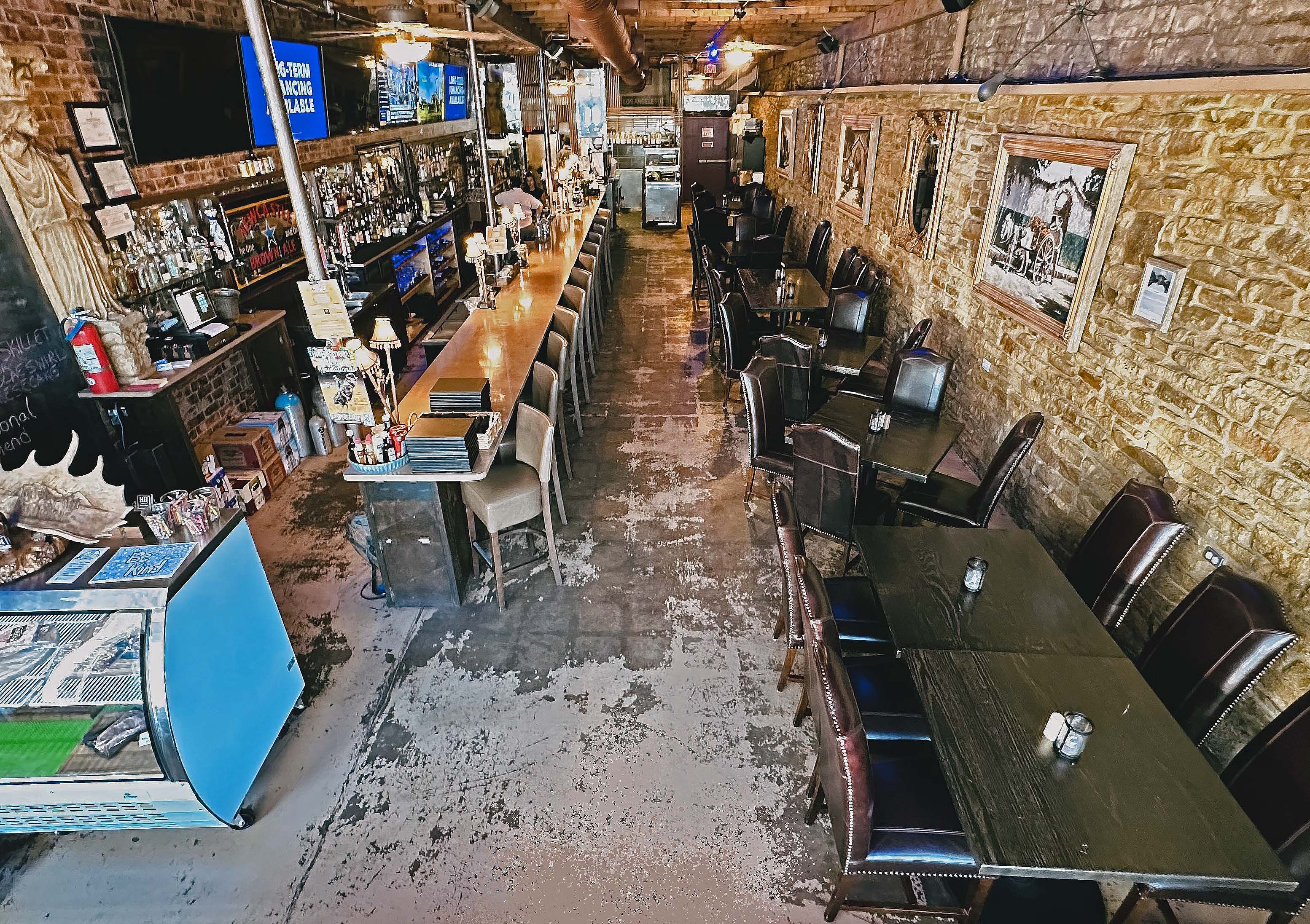 Interior of a rustic bar with a long wooden counter, bar stools, tables with chairs, exposed brick and stone walls decorated with framed pictures.