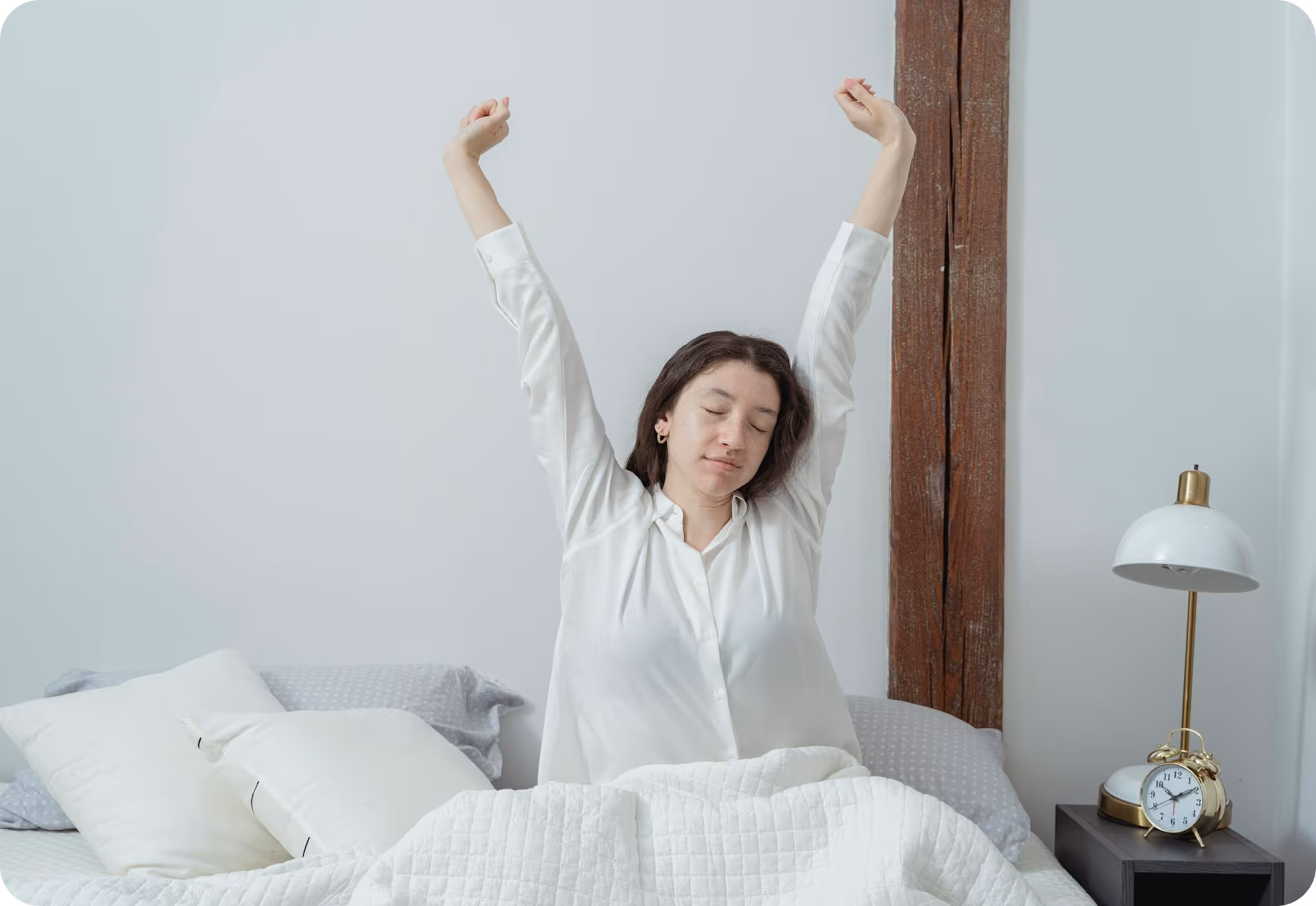 a woman sitting up in bed, stretching with her arms raised above her head. She has her eyes closed and appears to be waking up. She is wearing a white button-up shirt, and the bedding is white with some gray pillows. To the right, on a bedside table, there’s a white and gold desk lamp and a classic gold alarm clock. The background includes a white wall with a vertical wooden beam. The atmosphere is calm and cozy, evoking a morning routine scene.