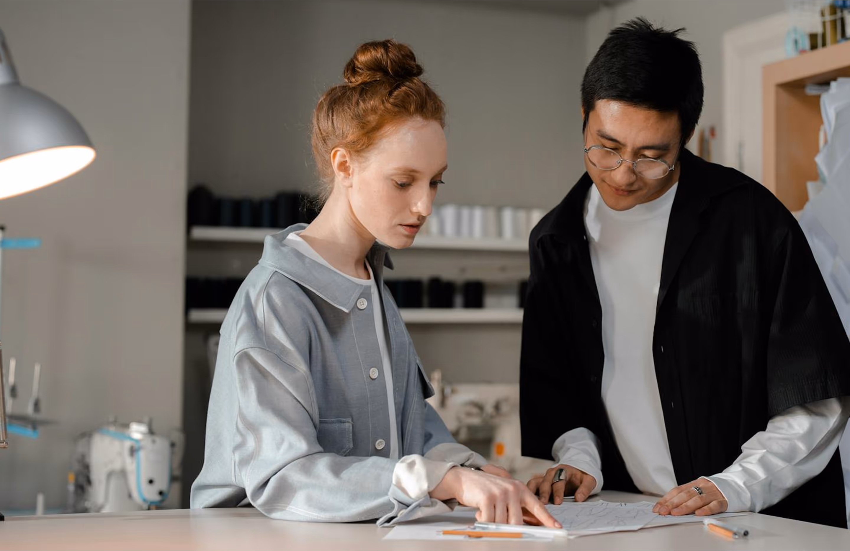 two people collaborating in a workspace, possibly a fashion or design studio. One person is pointing at a paper with sketches or technical drawings, while the other is leaning in to examine the details. There are sewing machines and spools of thread in the background, reinforcing the setting as a garment production or design environment.