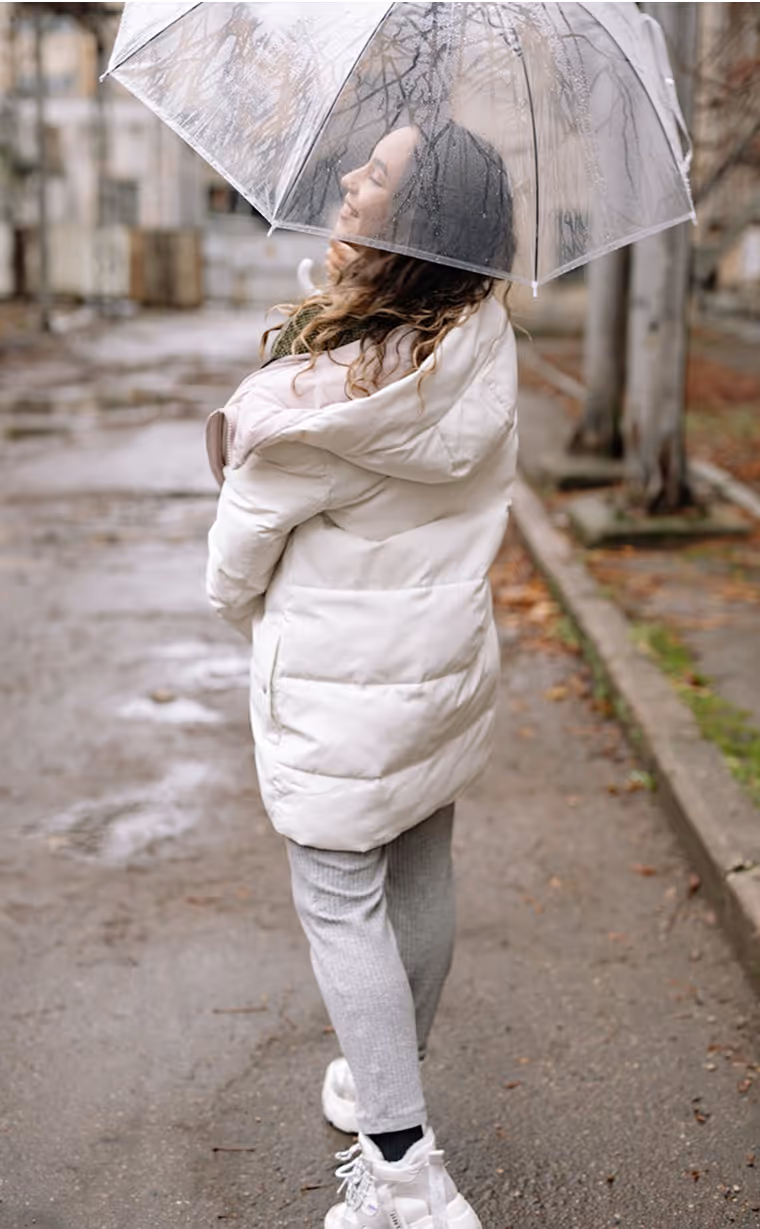 a woman walking outdoors on a rainy day, holding a transparent umbrella. She is wearing a white puffer jacket, gray pants, and white sneakers, smiling as she looks upward.