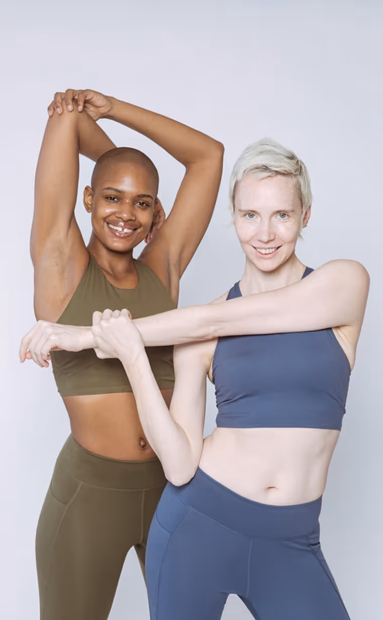 two women in activewear posing and stretching. The woman on the left is wearing an olive-green sports bra and matching leggings, while the woman on the right is wearing a blue sports bra and matching leggings. Both are smiling and standing against a light background.