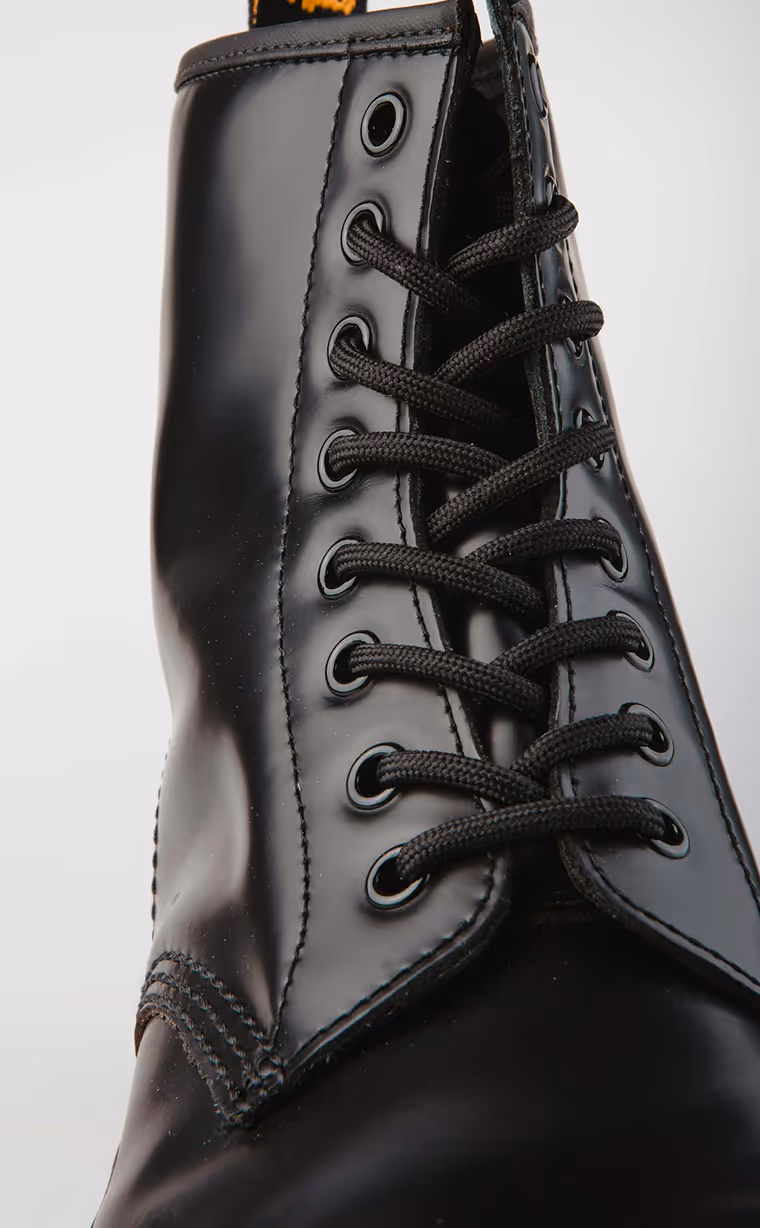 A close-up of a shiny black leather lace-up boot, featuring black laces threaded through metal eyelets. The top of the boot has a pull tab