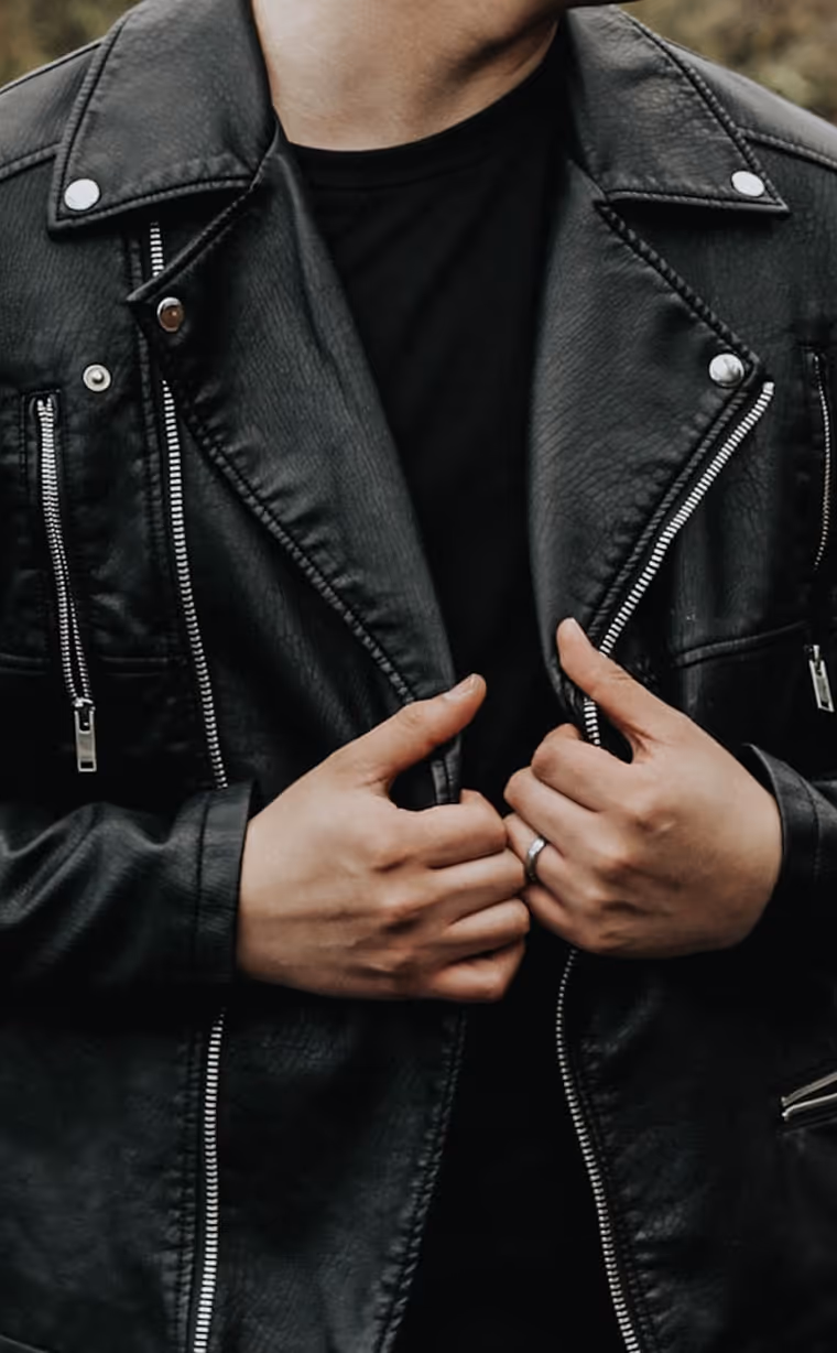 A person wearing a black leather jacket over a black shirt, holding the jacket edges with both hands. The jacket features silver zippers and snap buttons, and the person has a simple metallic ring on the ring finger of their right hand.