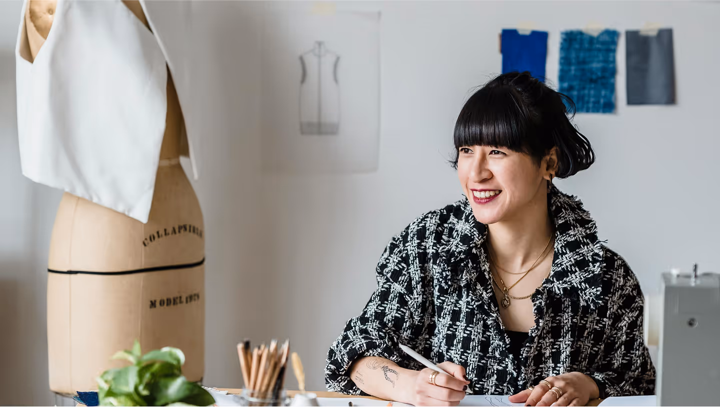 Smiling fashion designer sitting at a desk, sketching designs. She is wearing a black and white textured jacket and has black hair styled in a bun with bangs. In the background, there is a dress form with a partially constructed white garment, sketches and fabric swatches pinned to the wall, and various design tools on the desk.