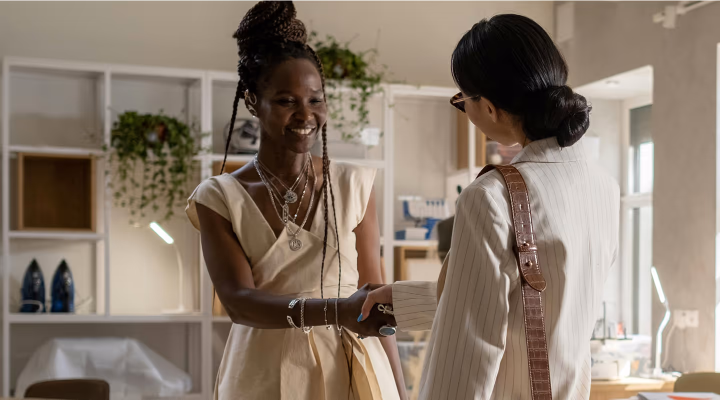 Two women shaking hands in a fashion design studio. The woman on the left, wearing a beige dress and layered necklaces, smiles warmly. The woman on the right, dressed in a light pinstriped suit with a brown shoulder strap, faces away from the camera. The background shows shelves with plants, irons, and sewing equipment.