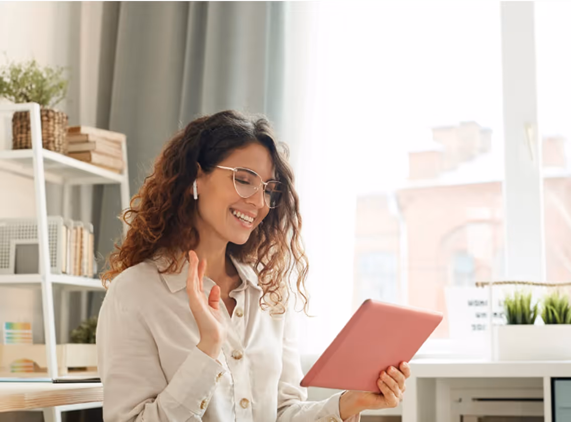 Smiling woman with curly hair and glasses, wearing a white shirt and wireless earbuds, holding a pink tablet and waving during a video call. She is seated in a bright, cozy room with a bookshelf, potted plant, and large window in the background.