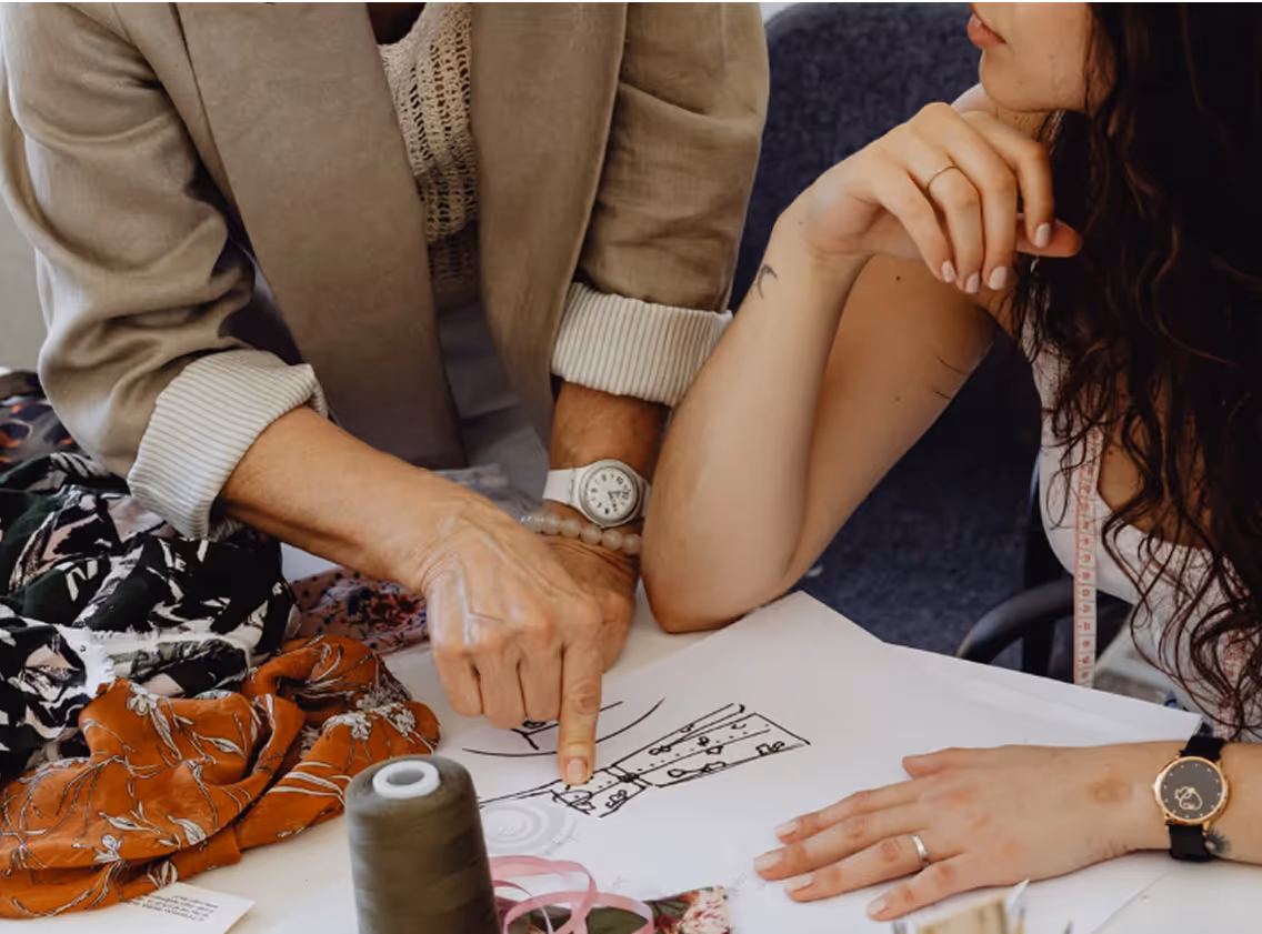 Close-up of two people collaborating on a fashion design project. One person wearing a beige blazer points to a sketch of a dress on paper, while the other person with long curly hair and a measuring tape around their neck listens attentively. The table is covered with patterned fabrics, a spool of green thread, and various sewing materials.
