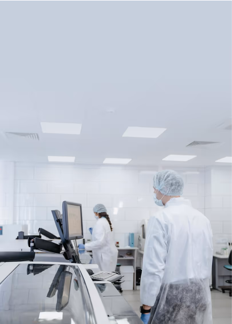 Two laboratory technicians wearing protective gowns, hairnets, face masks, and gloves working in a bright, modern laboratory. One technician is standing near a computer workstation while the other moves between lab equipment in the background. The lab has white tiled walls, overhead fluorescent lighting, and various scientific instruments.