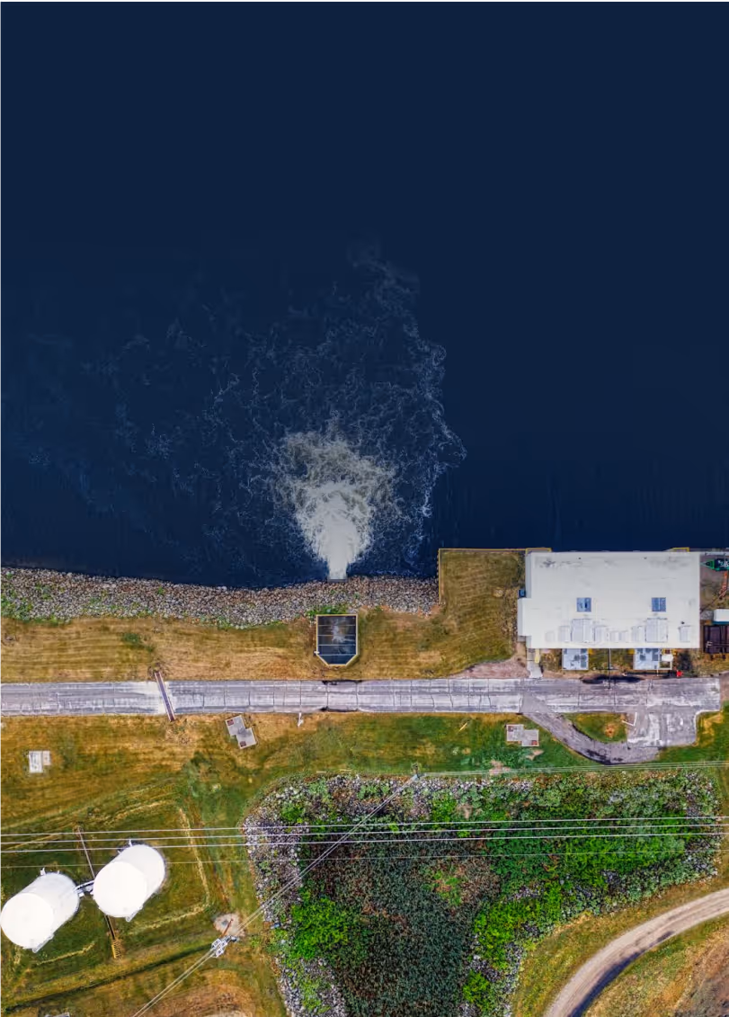 Aerial view of an industrial facility discharging water into a large dark body of water, creating visible turbulence at the outlet. The shoreline is lined with rocks, and the facility building is adjacent to a paved road. The surrounding area includes grassy fields, two large white storage tanks, electrical lines, and patches of vegetation.