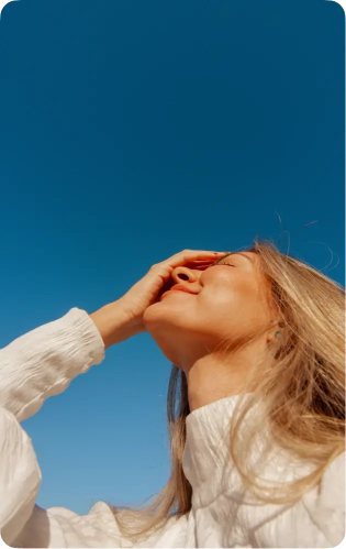 Woman with long blonde hair wearing a white sweater, smiling with eyes closed against a clear blue sky, holding one hand over her forehead.