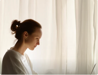Woman with hair in a bun wearing a white sweater sitting and looking down thoughtfully against a light curtain background.