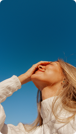 Woman with long blonde hair wearing a white sweater, smiling with eyes closed against a clear blue sky, holding one hand over her forehead.