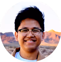 A young man with black hair and glasses is smiling at the camera. He is wearing a white shirt and a tan jacket. The background is a desert landscape with mountains under a clear sky.