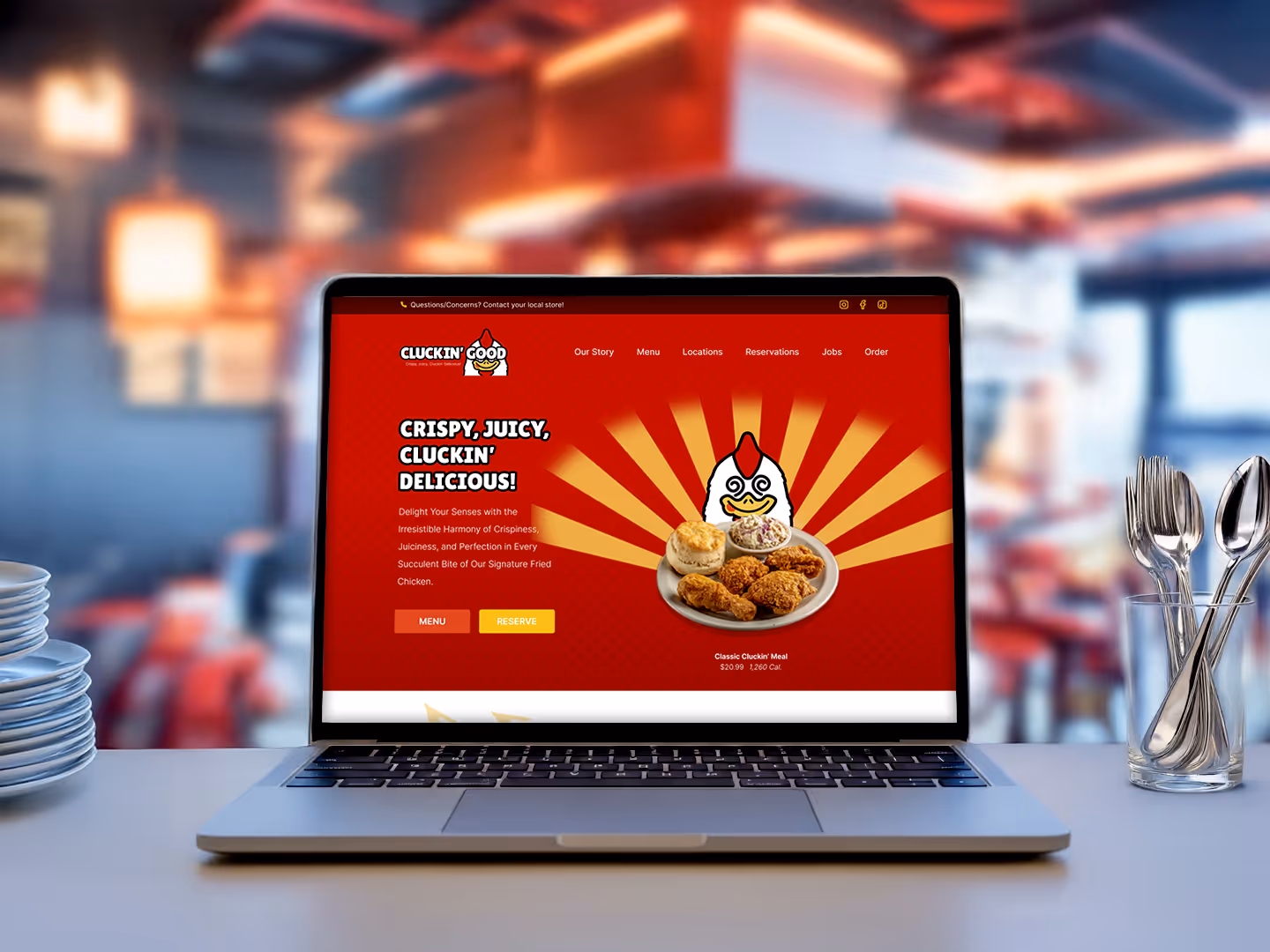 A laptop sits on a table in a restaurant, displaying the homepage of the "Cluckin' Good" fried chicken restaurant website. The site features a red background, a cartoon chicken mascot, and an image of a fried chicken meal with a biscuit.