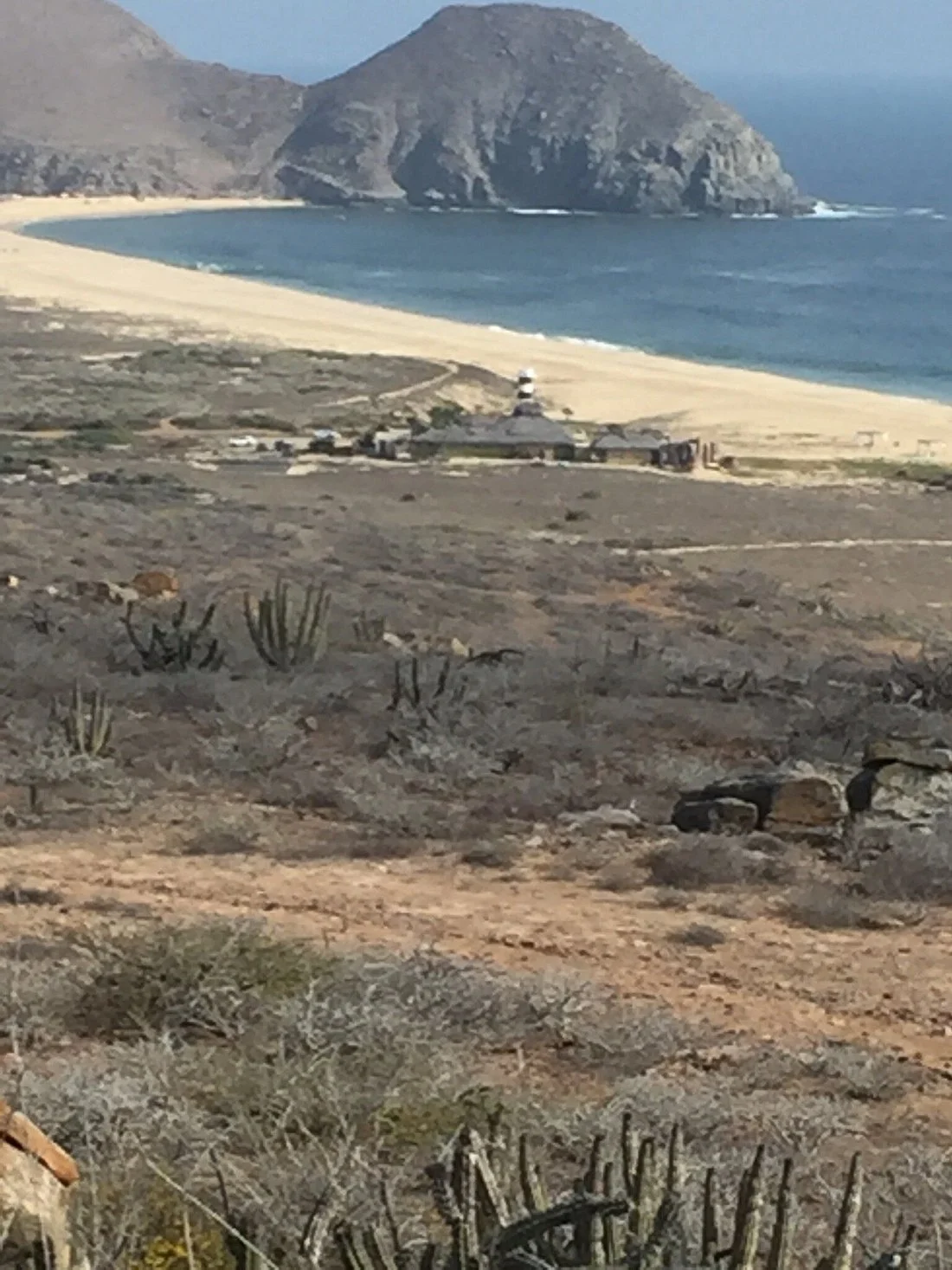 Vista de una playa extensa con arena clara, un faro cerca de la costa y montañas rocosas al fondo, rodeada de vegetación desértica Guaycura Hotel Todos Santos