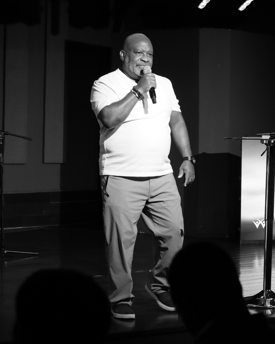 Man in a white shirt holding a microphone speaking on stage in front of an audience.