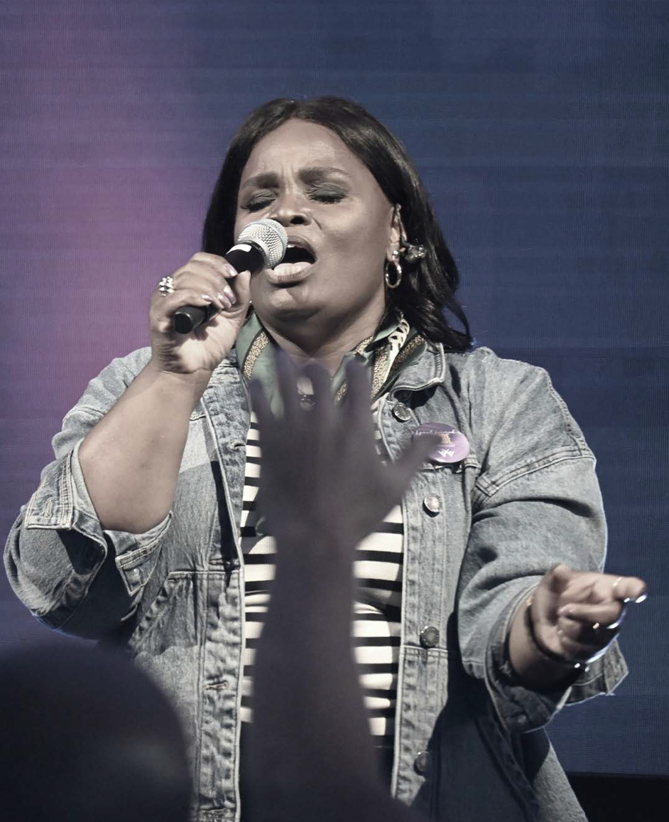 Female singer passionately singing into a microphone with audience hand raised in foreground.