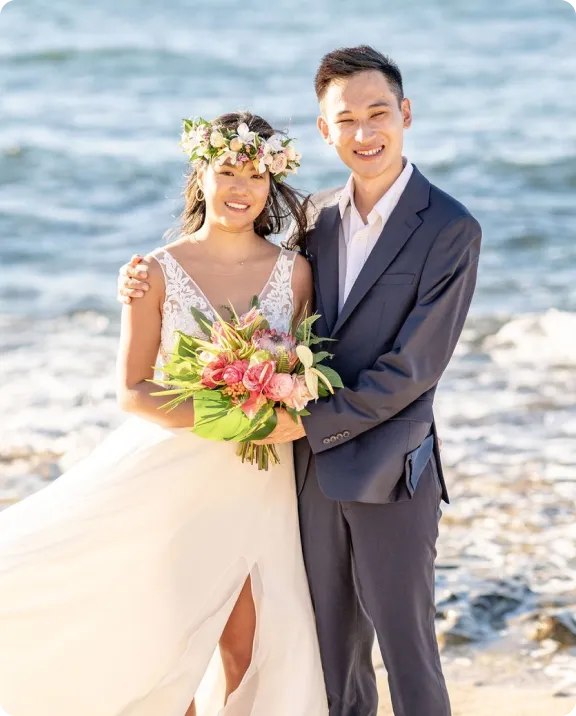 Couple in wedding attire standing on a beach with waves behind them.