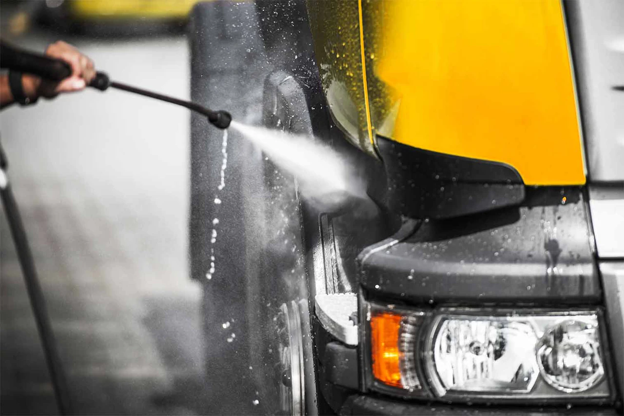 A person uses a high-pressure washer to clean the front tire and fender of a yellow and black semi-truck. Water sprays forcefully, creating mist and reflections on the wet truck surface.
