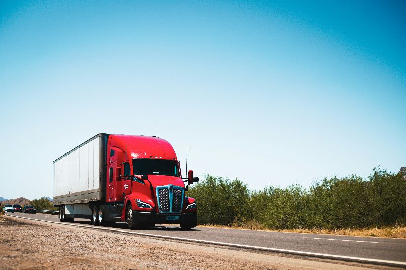 Bright red semi truck pulling a box trailer down an open highway bordered by desert brush beneath a clear sunny sky.