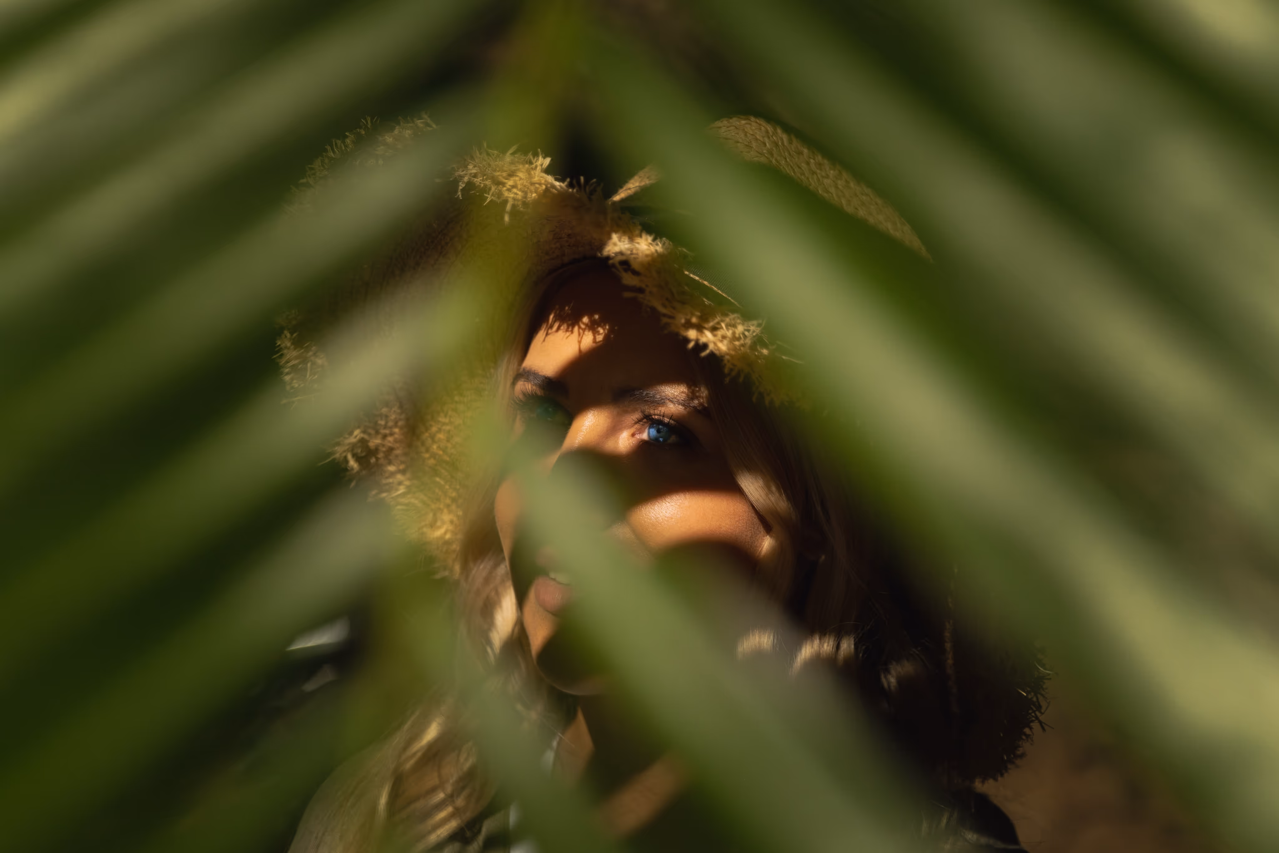 Mysterious portrait of a woman in a straw hat, partially hidden behind green palm leaves, with dramatic sunlight highlighting her blue eyes and face. The artistic composition evokes themes of nature, curiosity, and tropical adventure photography.