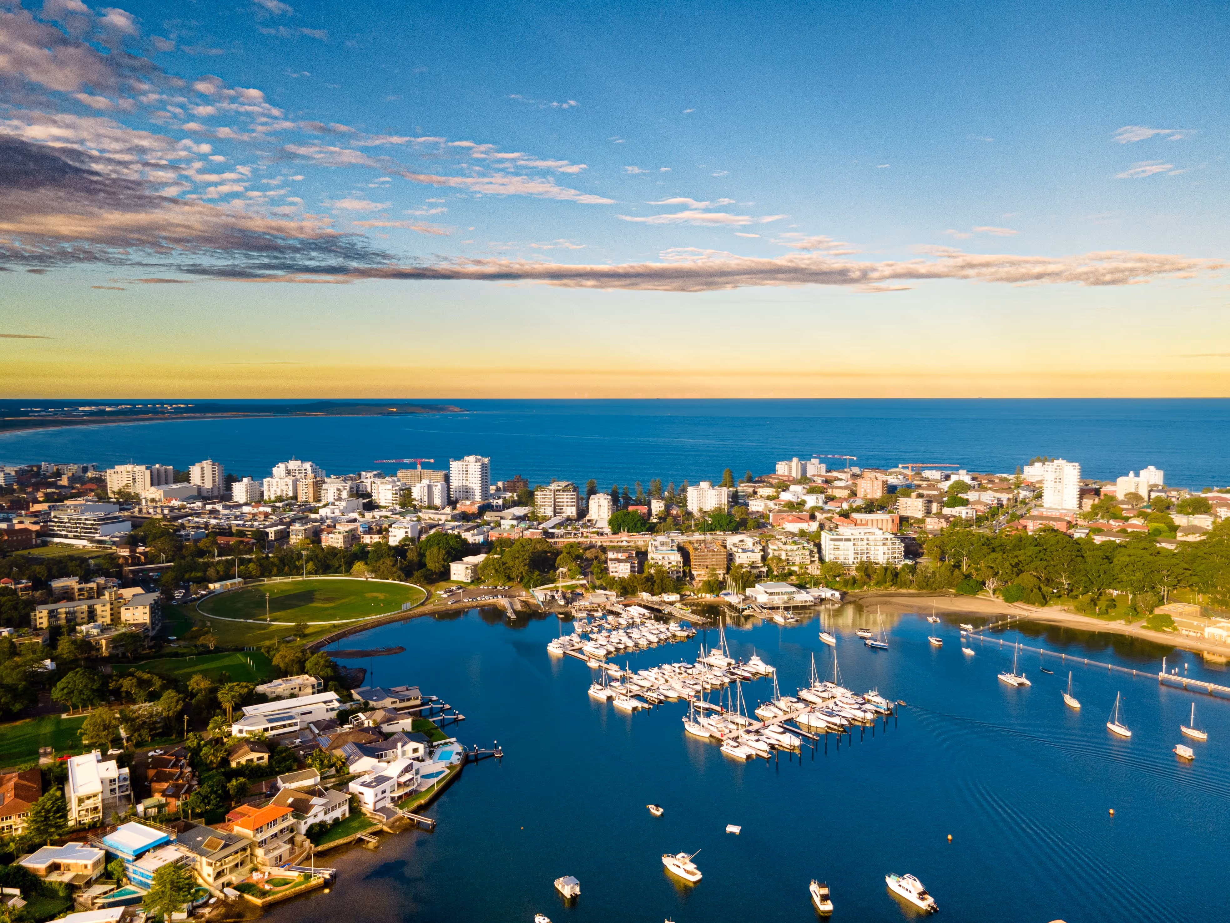 Aerial view of a coastal city at sunrise, featuring a vibrant marina filled with yachts and boats, residential areas with waterfront homes, and a skyline of modern apartment buildings. The calm ocean and golden sky create a serene setting ideal for tourism, luxury real estate, or coastal lifestyle content.