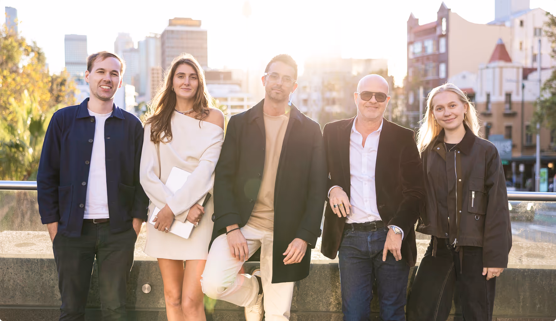 Group of five stylish professionals standing outdoors in front of a city skyline during golden hour, smiling and casually posed. Ideal for themes like modern business team, startup culture, creative agency branding, or urban professional lifestyle.