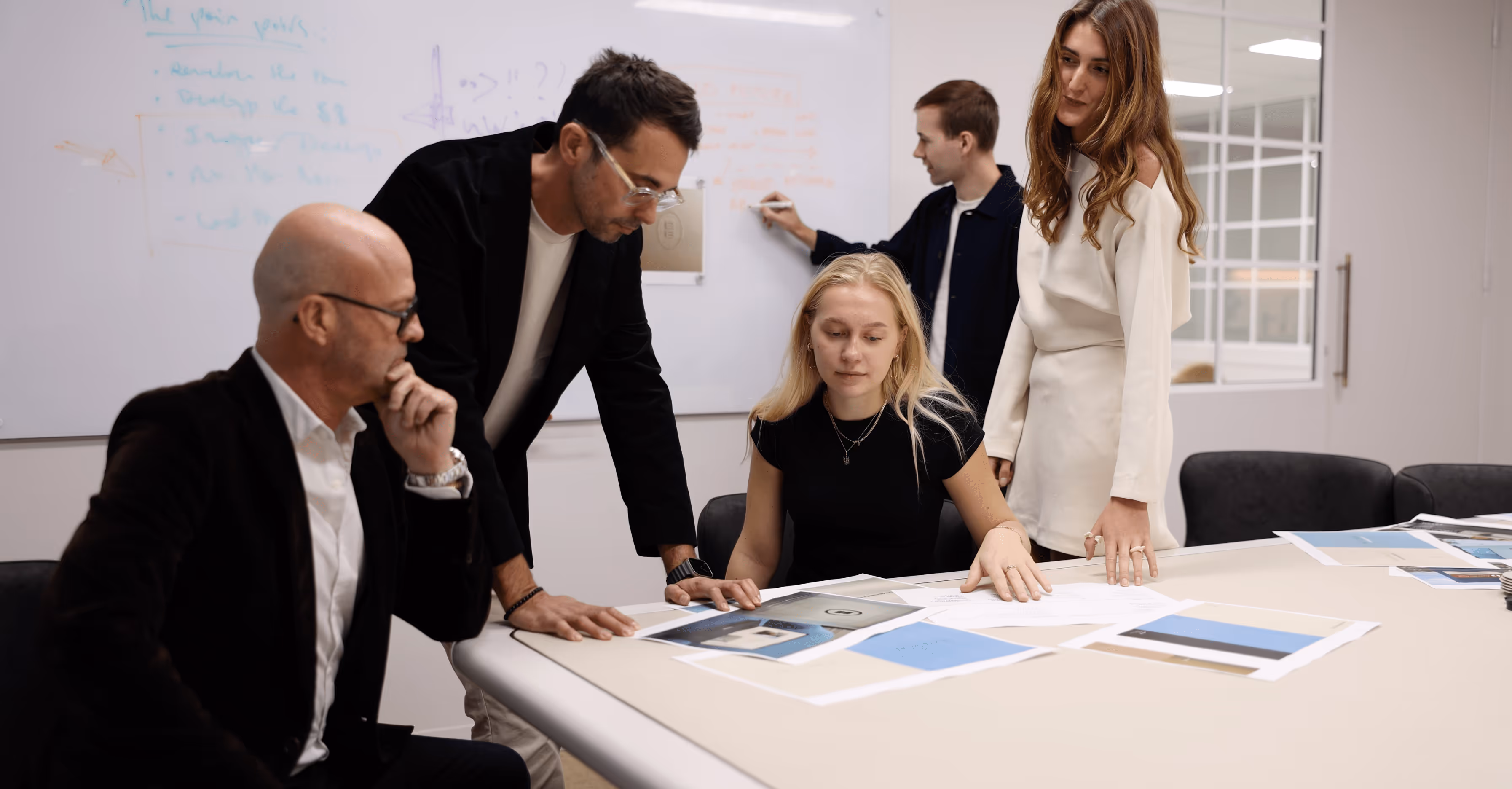 Creative team in a modern office collaborating on a branding or design project, reviewing printed materials around a conference table. One team member writes on a whiteboard while others engage in discussion, reflecting teamwork, visual strategy, and project planning.