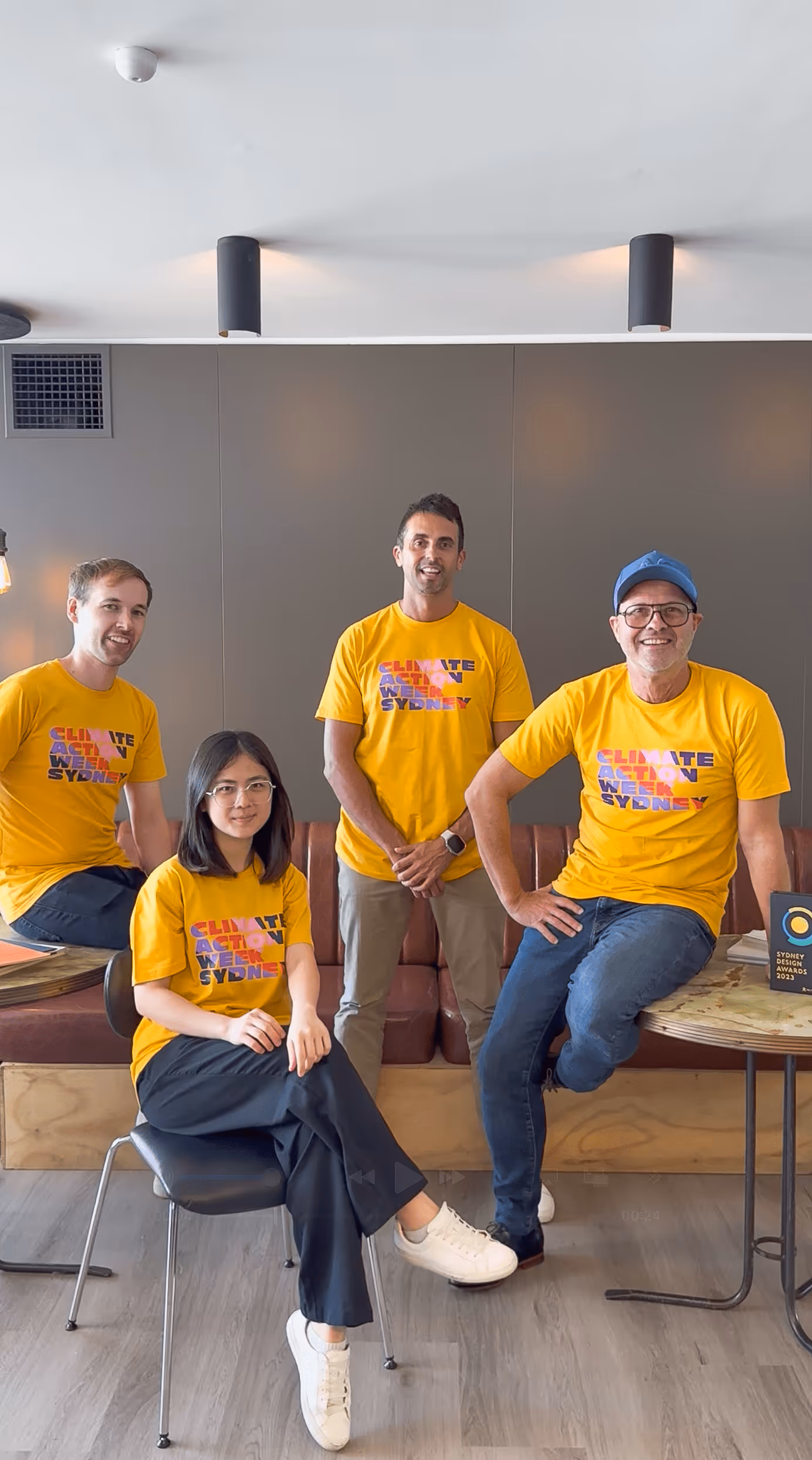 Four team members wearing bright yellow "Climate Action Week Sydney" t-shirts pose together indoors, smiling and relaxed. The group is part of a sustainability or environmental event, with a Sydney Design Awards 2023 trophy visible on a nearby table.