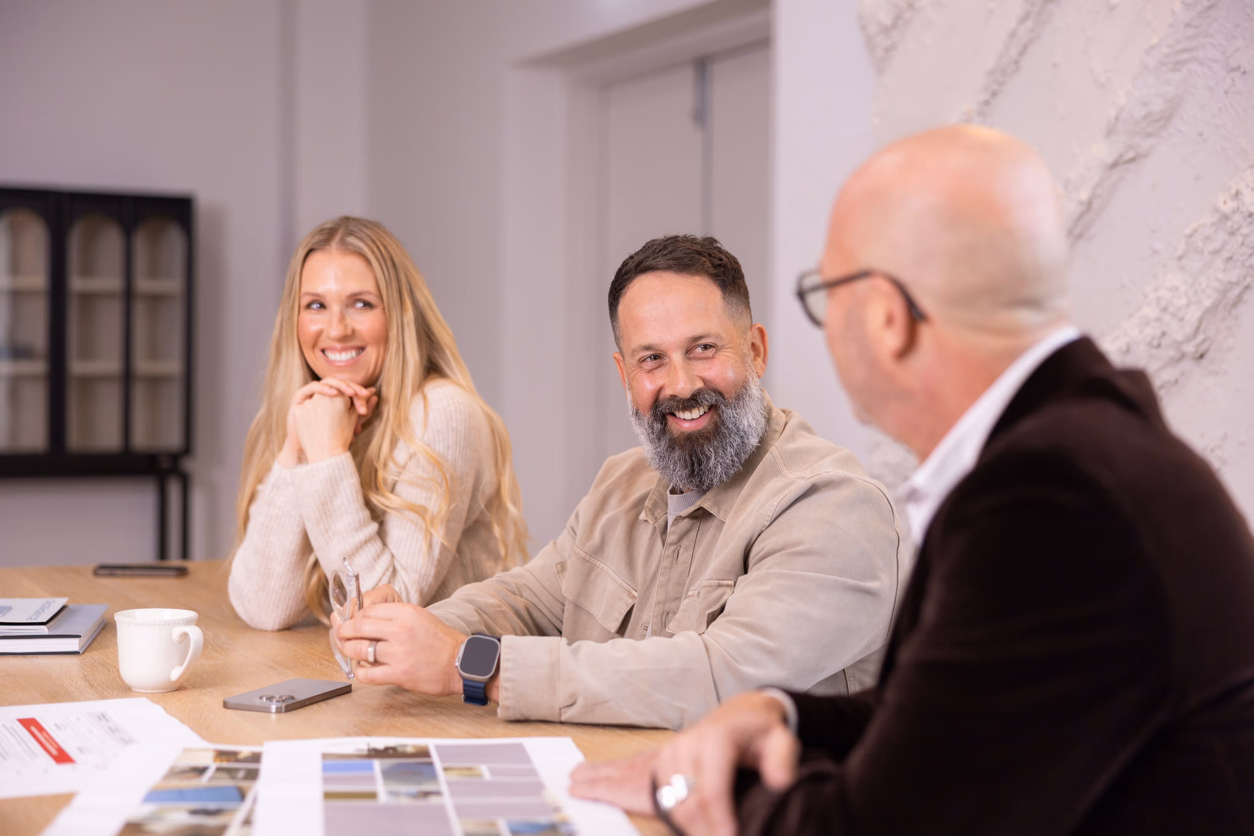 Three professionals engaged in a casual and friendly business meeting, smiling and discussing project materials at a modern office table. The scene reflects creative collaboration, client consultation, and relaxed team communication.