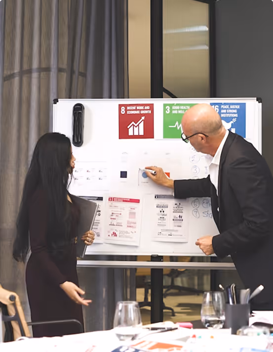 Two professionals collaborating at a whiteboard covered in charts and UN Sustainable Development Goals posters, including Goal 8 (Decent Work), Goal 3 (Good Health), and Goal 16 (Peace and Justice). The scene represents strategic planning, sustainability consulting, and corporate responsibility initiatives.